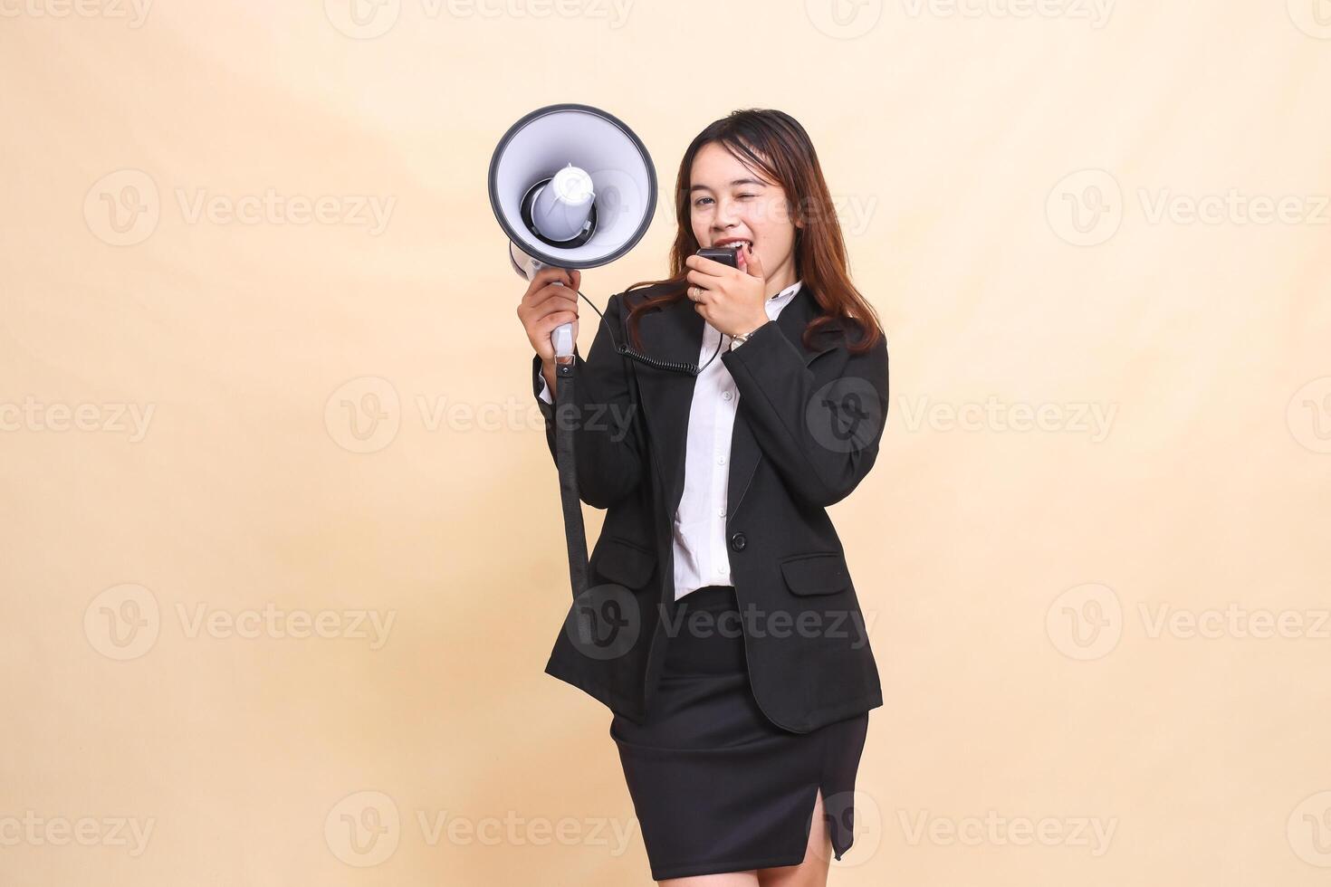 Beautiful formal Asian office woman in suit standing happy shouting winking at camera with mic using megaphone for promo broadcast announcement on yellow background photo