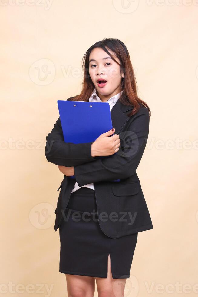 Young beautiful formal Asian executive woman in suit shocked standing and pose hugging pen clipboard to camera, 3 by 4 body portrait on yellow background for commercial, vacancy and business photo