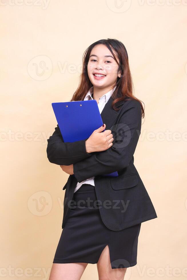 Young beautiful formal Asian executive woman in suit cheerful smile standing and pose hugging pen clipboard to camera, 3 by 4 body portrait on yellow background for commercial, vacancy and business photo