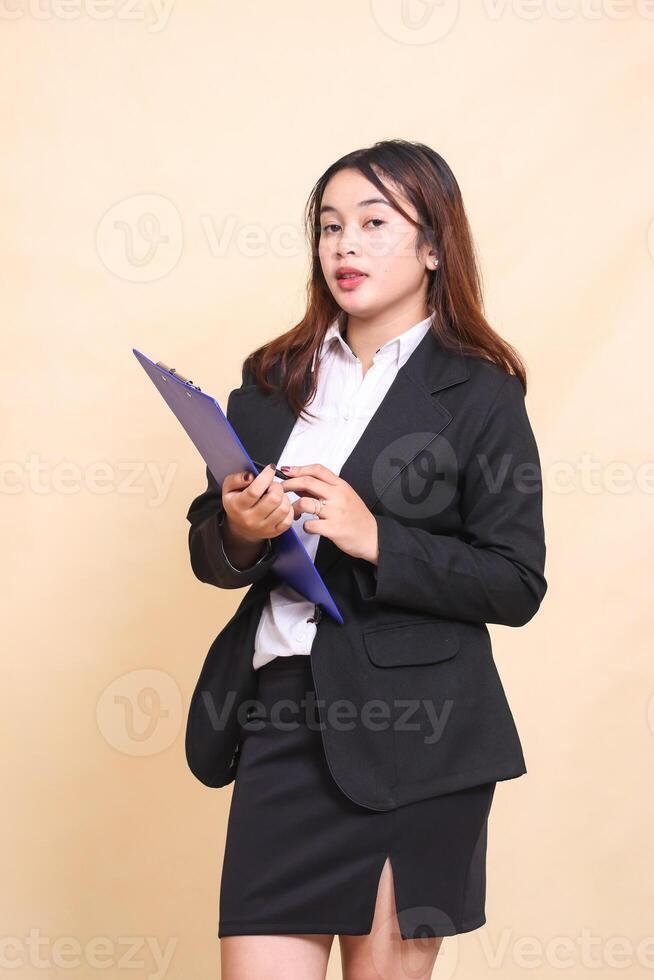 Beautiful formal Asian executive mature woman in cool suit standing and posing holding clipboard pen to camera, 3 by 4 body portrait on yellow background for commercial, vacancy and corporate photo