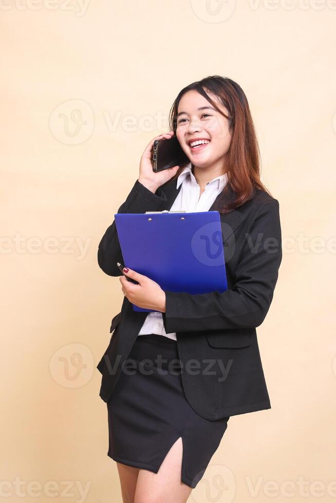 Beautiful formal Asian executive girl in suit standing happy and pose hugging clipboard and telephone with smartphone gadget candid on yellow background for commercial, vacancy and business photo