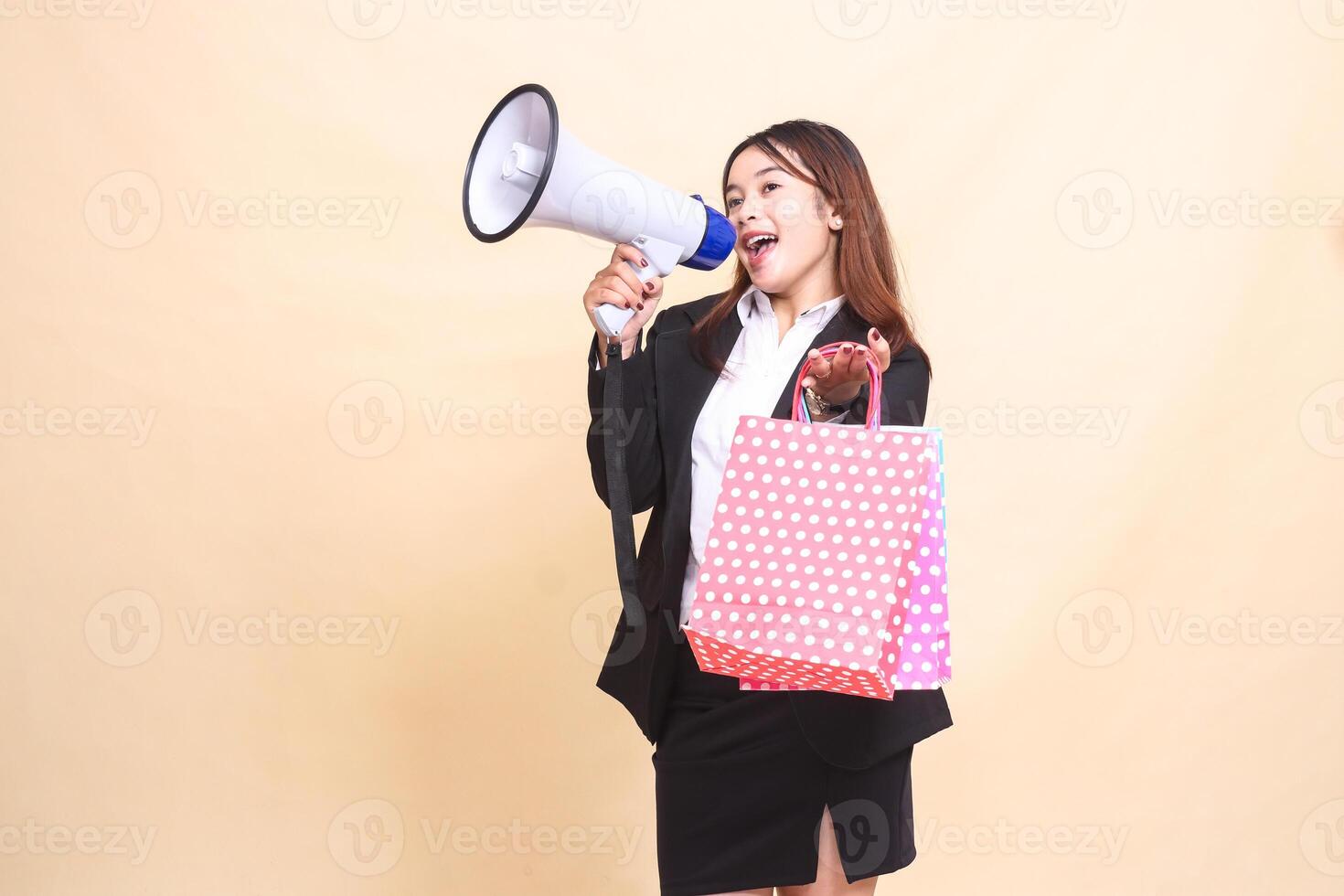 Beautiful formal Asian office girl in suit standing smiling shouting candid carrying shopping paperbag in front while carrying mic using megaphone for promo announcement on yellow background photo