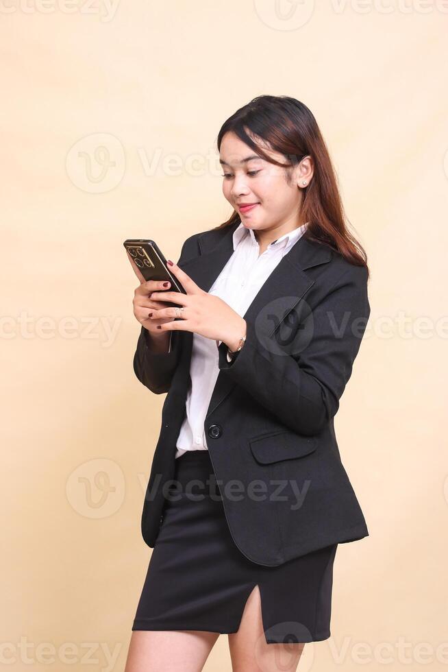 Beautiful formal Asian executive woman in suit standing happy smile and holding while operating online shopping gadget smartphone on yellow background for commercial, vacancy and business photo