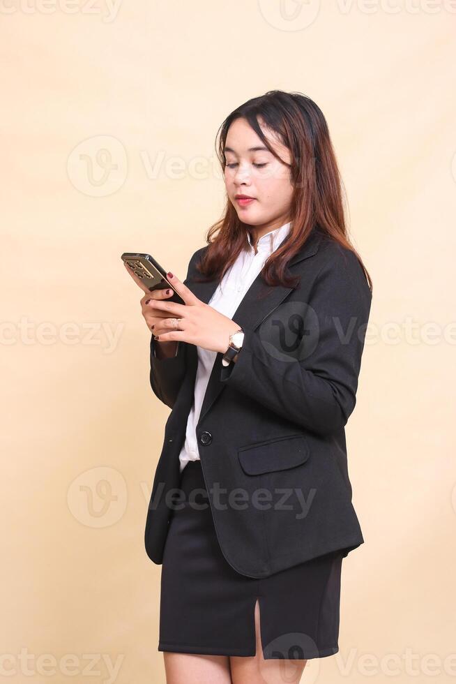 Beautiful formal Asian executive woman in suit standing and holding while operating smartphone gadget chatting with her friend on yellow background for commercial, vacancy and business photo