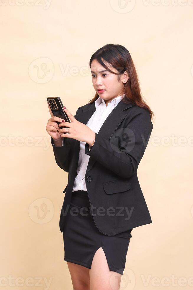 Beautiful formal Asian executive woman in suit standing serious expression while operating online shopping gadget smartphone on yellow background for commercial, vacancy and business photo