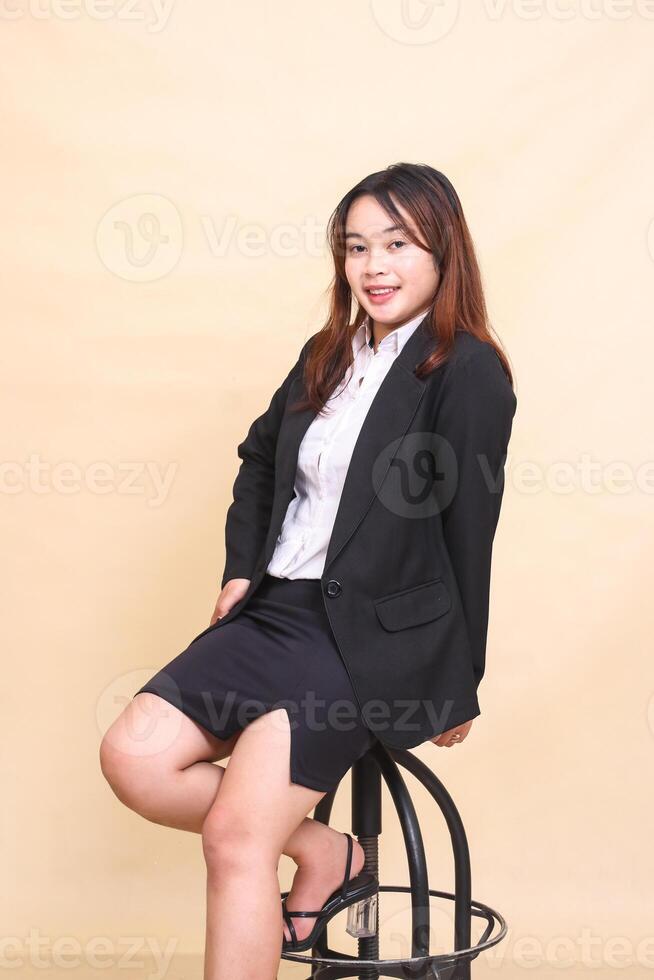 Young beautiful Asian executive woman in suit cheerful smile sitting on chair and pose holding chair to camera, 3 by 4 body portrait on yellow background for commercial, vacancy and corporate photo