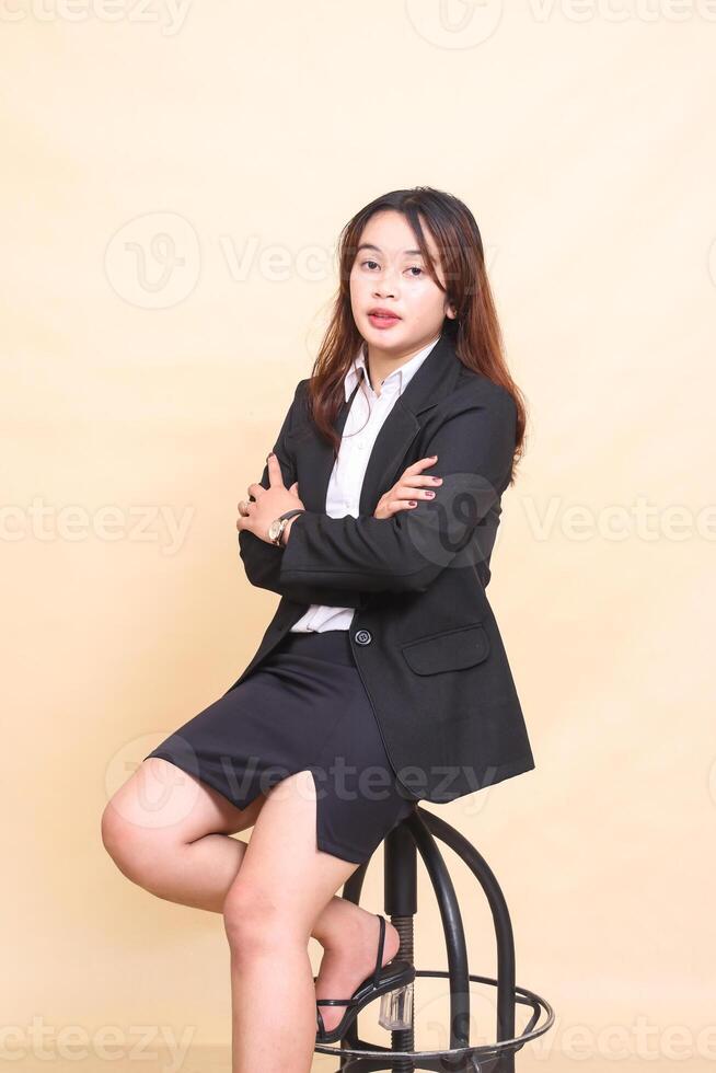 Young beautiful formal Asian executive woman in elegant suit sitting on chair and pose with arms to camera, 3 by 4 body portrait on yellow background for commercial, vacancy and business photo