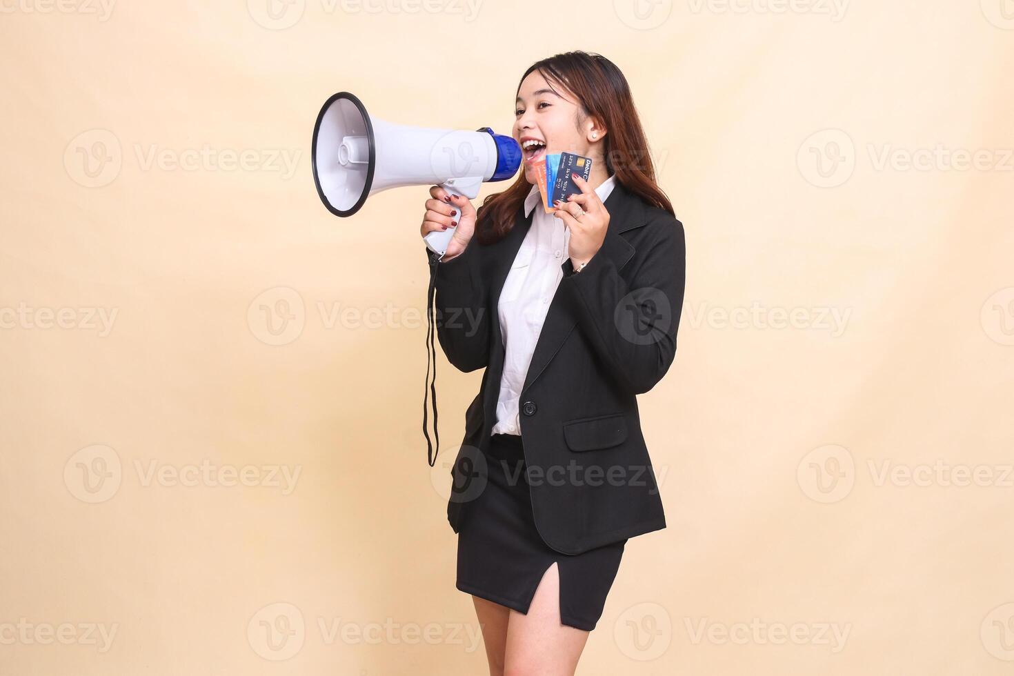 Beautiful formal Asian office woman in suit standing screaming holding debit credit card and megaphone for broadcast announcement on yellow background for commercial, vacancy and business photo