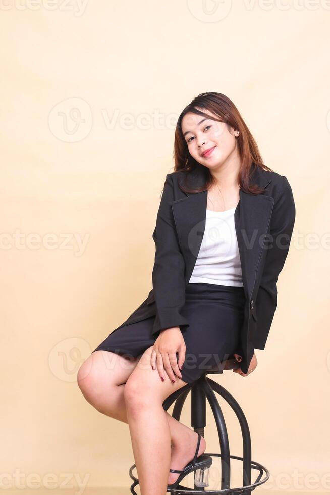 Young beautiful Asian executive woman smile sitting on chair and pose holding thighs and chair to camera, 3 by 4 body portrait on yellow background for commercial, vacancy and business photo