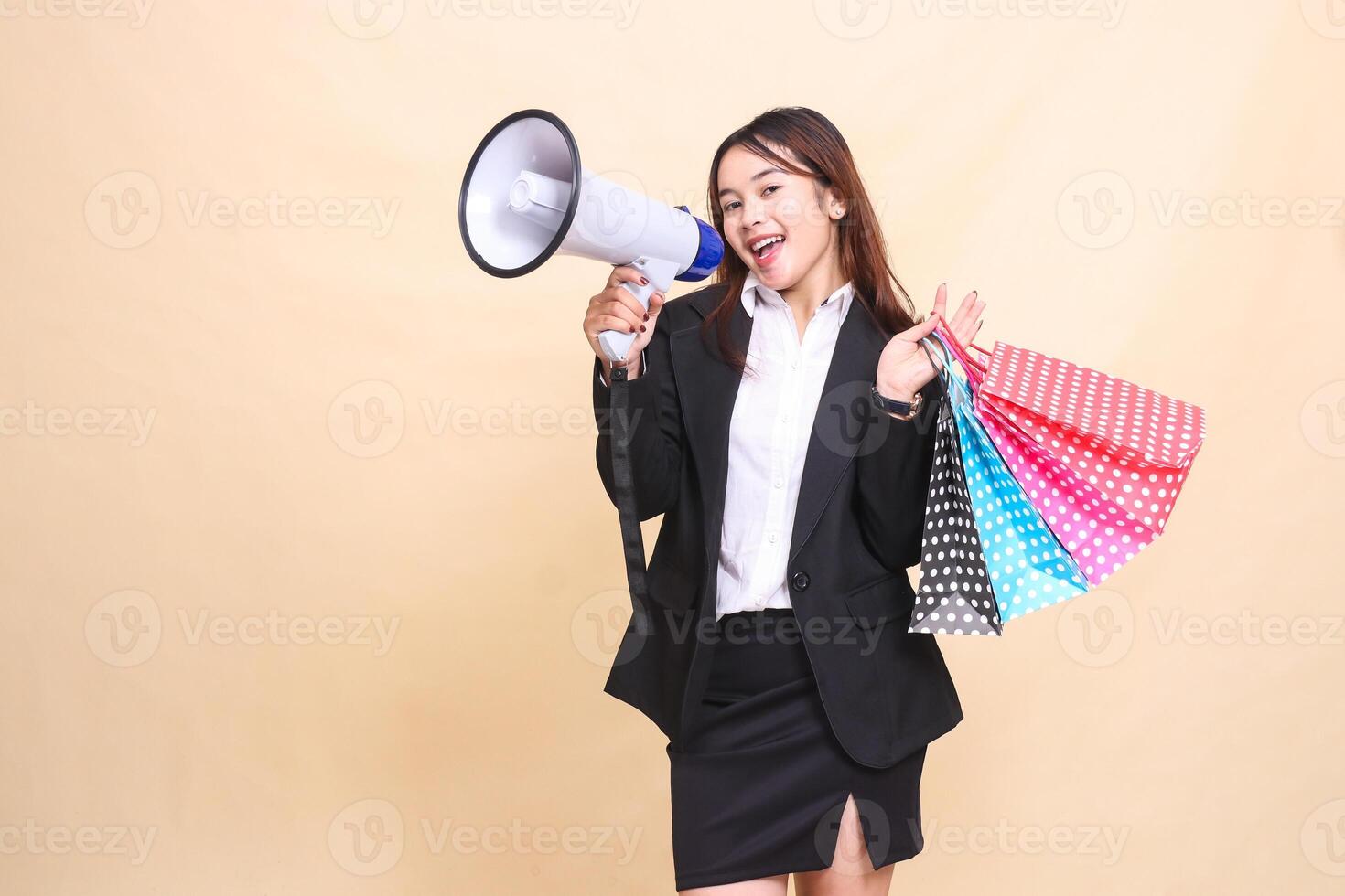Beautiful formal Asian office woman in suit standing smiling shouting at camera lifting shopping paperbag while carrying mic using megaphone for promo announcement on yellow background photo