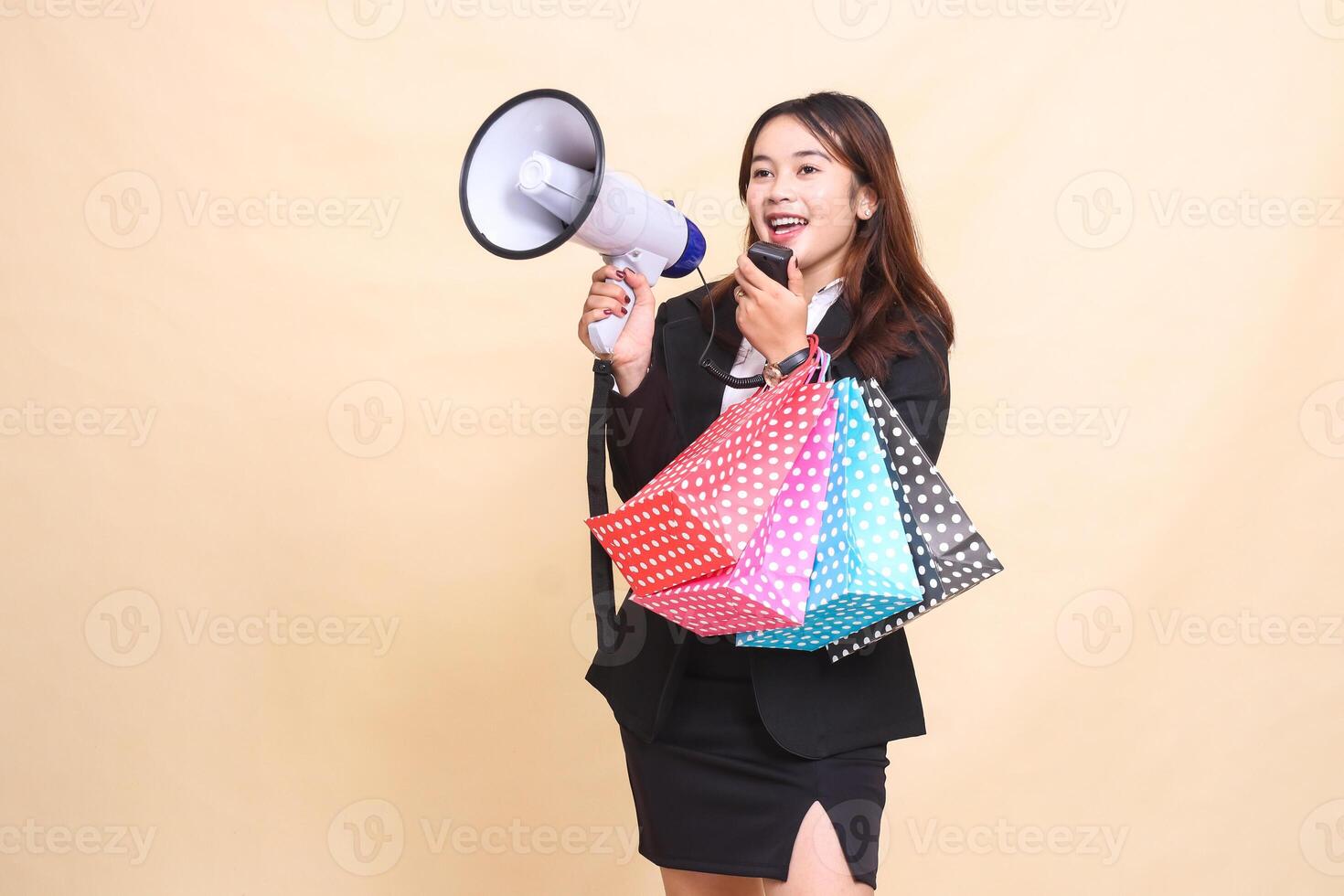 Beautiful formal Asian office woman in suit standing carrying shopping paperbag while carrying mic using megaphone for announcement promo on yellow background for commercial, advertising and business photo