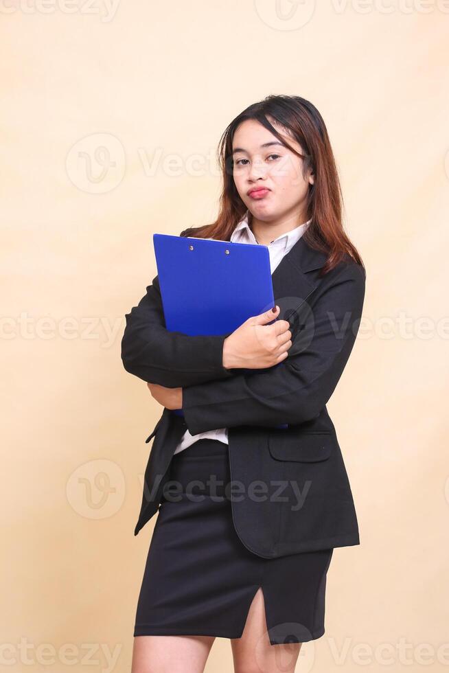 Young beautiful formal indonesia executive woman in suit frowning standing and posing hugging pen clipboard to camera, 3 by 4 body portrait on yellow background for commercial, vacancy and business photo
