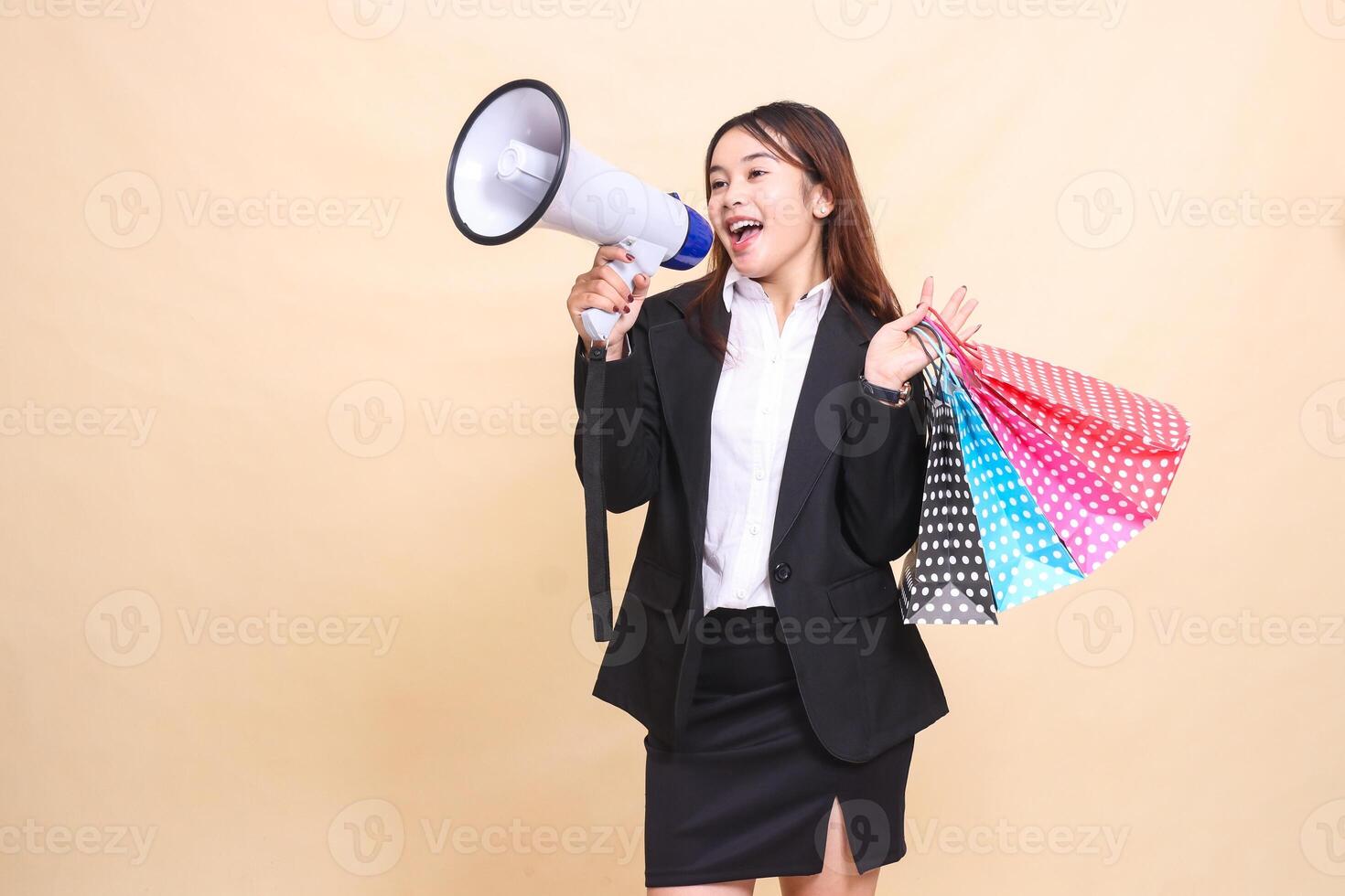 Beautiful formal Asian office woman in suit standing smiling shouting lifting shopping paperbag while carrying mic using megaphone for promo announcement on yellow background for commercial photo
