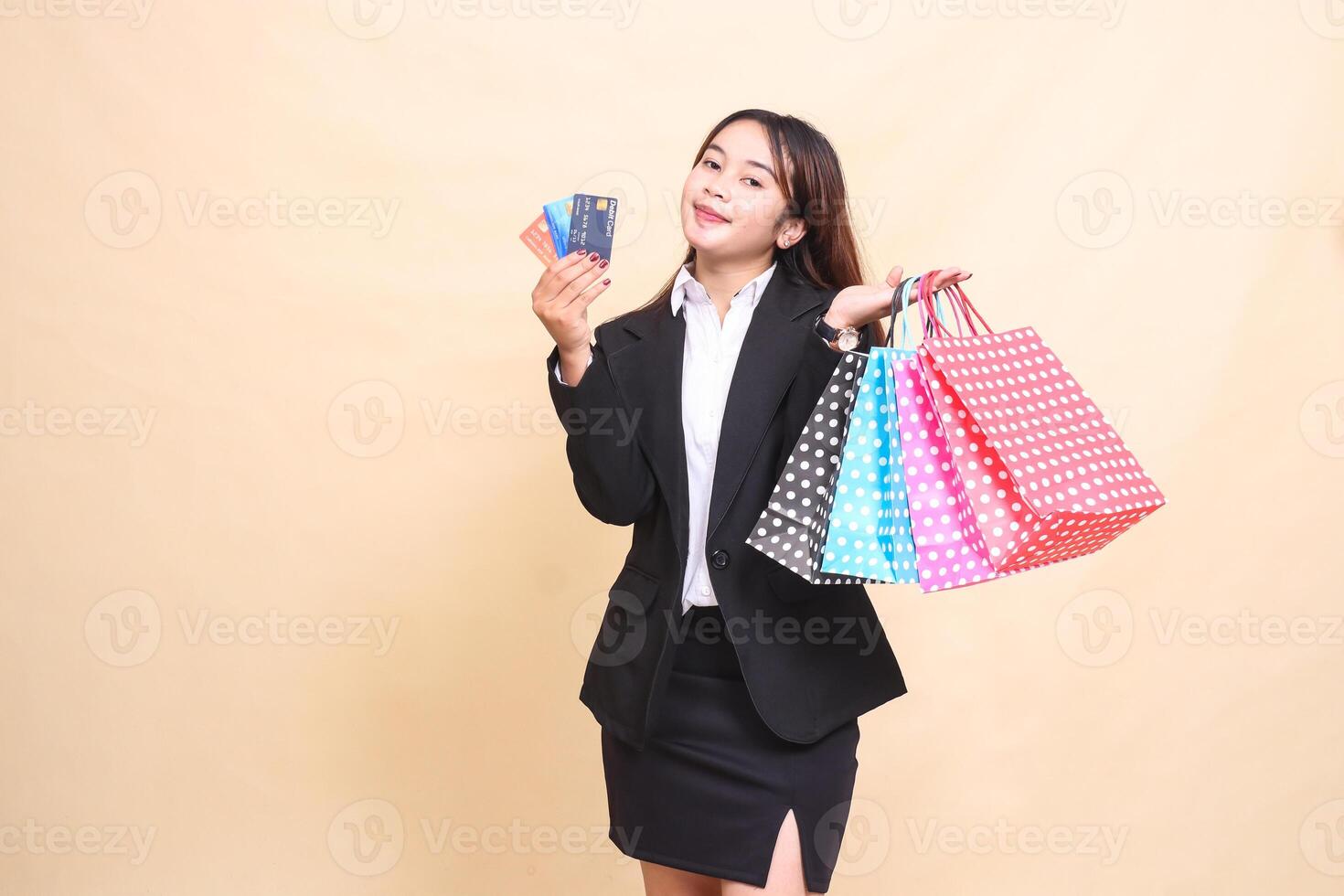 Beautiful formal Asian office woman in suit standing smiling carrying paper bag and credit and debit cards to start online shopping on yellow background for commercial, advertising and business photo