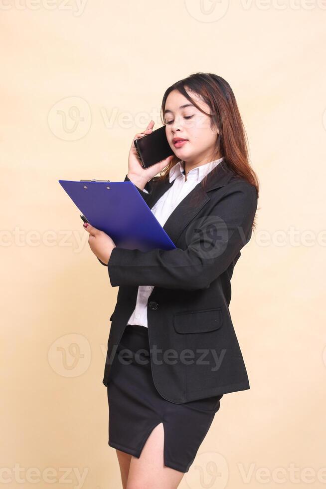 Beautiful formal indonesia executive woman in suit elegant standing and pose holding clipboard and telephone with smartphone gadget on yellow background for commercial, vacancy and business photo