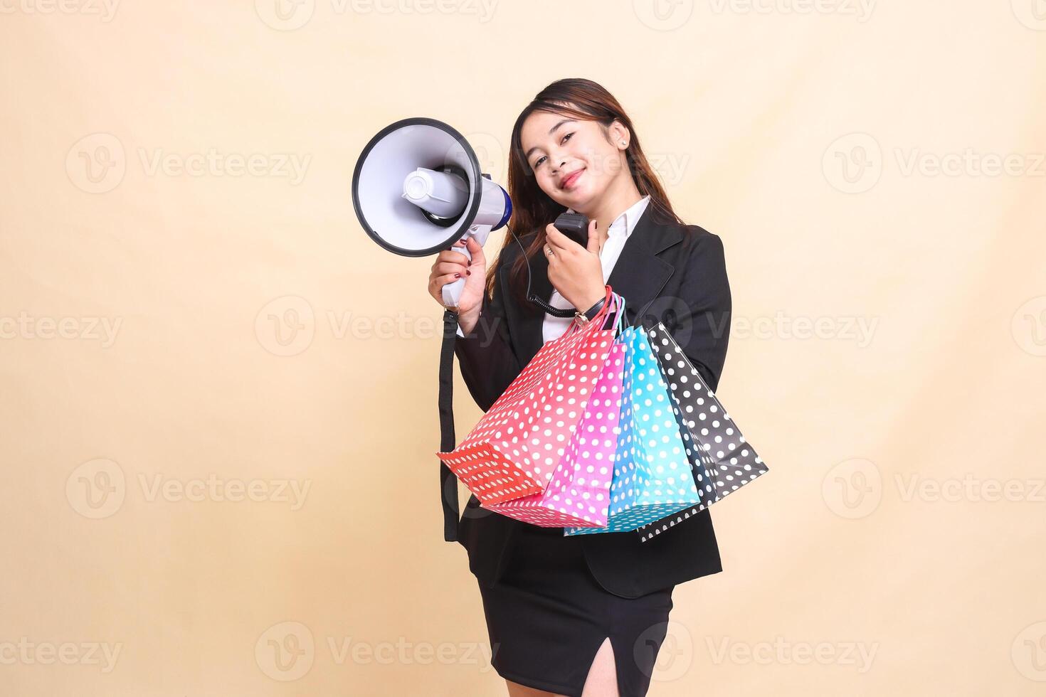 Beautiful formal Asian office woman in suit standing smiling carrying shopping paperbag while carrying mic using megaphone for promo announcement on yellow background for advertising and business photo