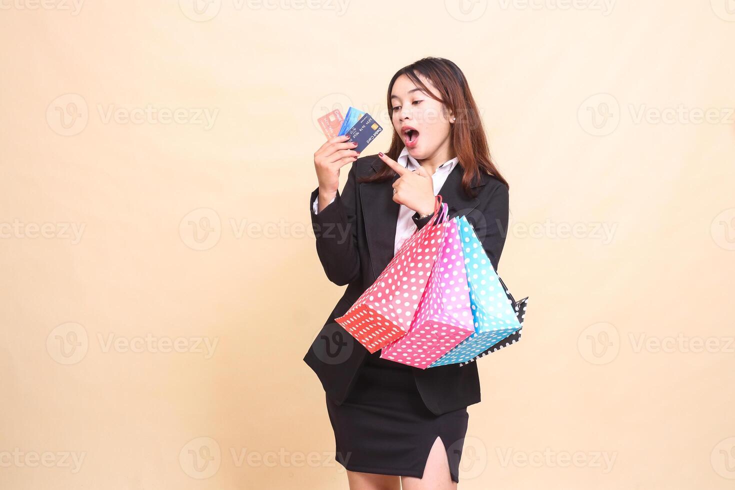 Beautiful formal indonesia office woman in suit standing surprised candid and carrying paper bag while pointing at debit credit card to start paying online on yellow background for commercial photo