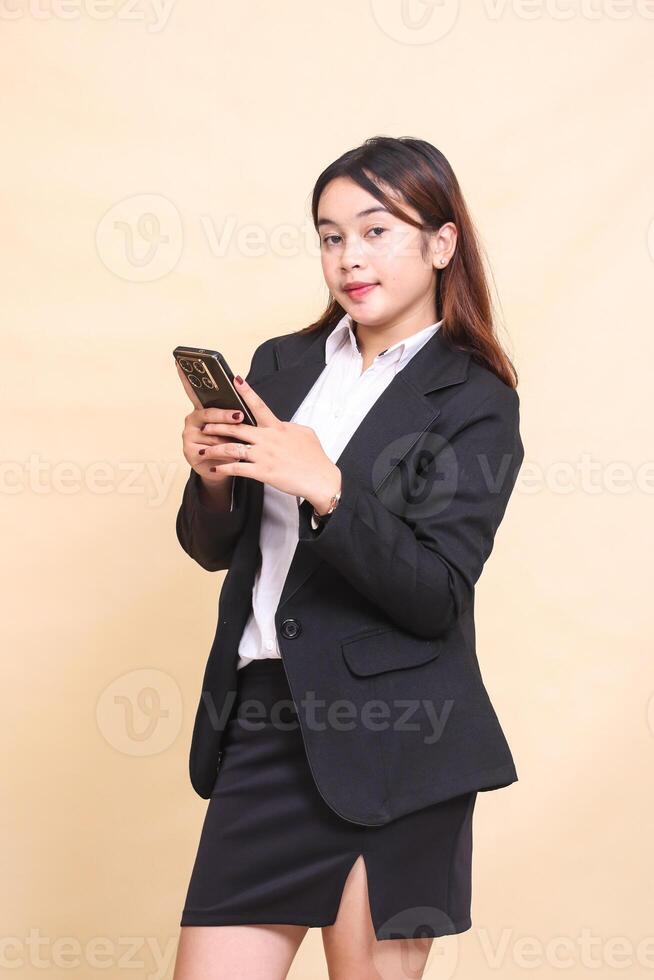 Beautiful formal indonesia executive woman in suit elegant standing and holding while operating smartphone gadget chatting with her friend on yellow background for commercial, vacancies and business photo