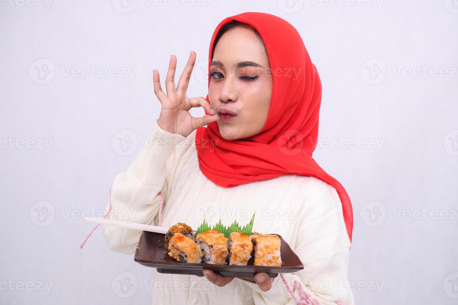 Asian woman in hijab kissing winks at the camera with a delicious gesture and both hands hold a plate containing sushi Japanese food. Beautiful Muslim women use culinary, food and lifestyle content photo