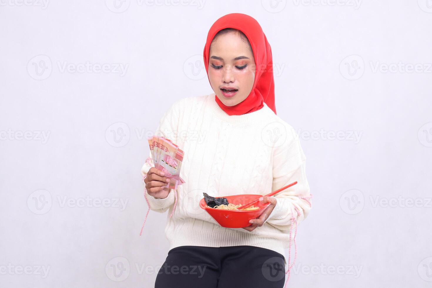 Asian woman wearing a hijab is shocked to see ramen holding rupiah notes and a bowl containing ramen Chinese food and chopsticks. Beautiful Muslim women are used for culinary and advertising content photo