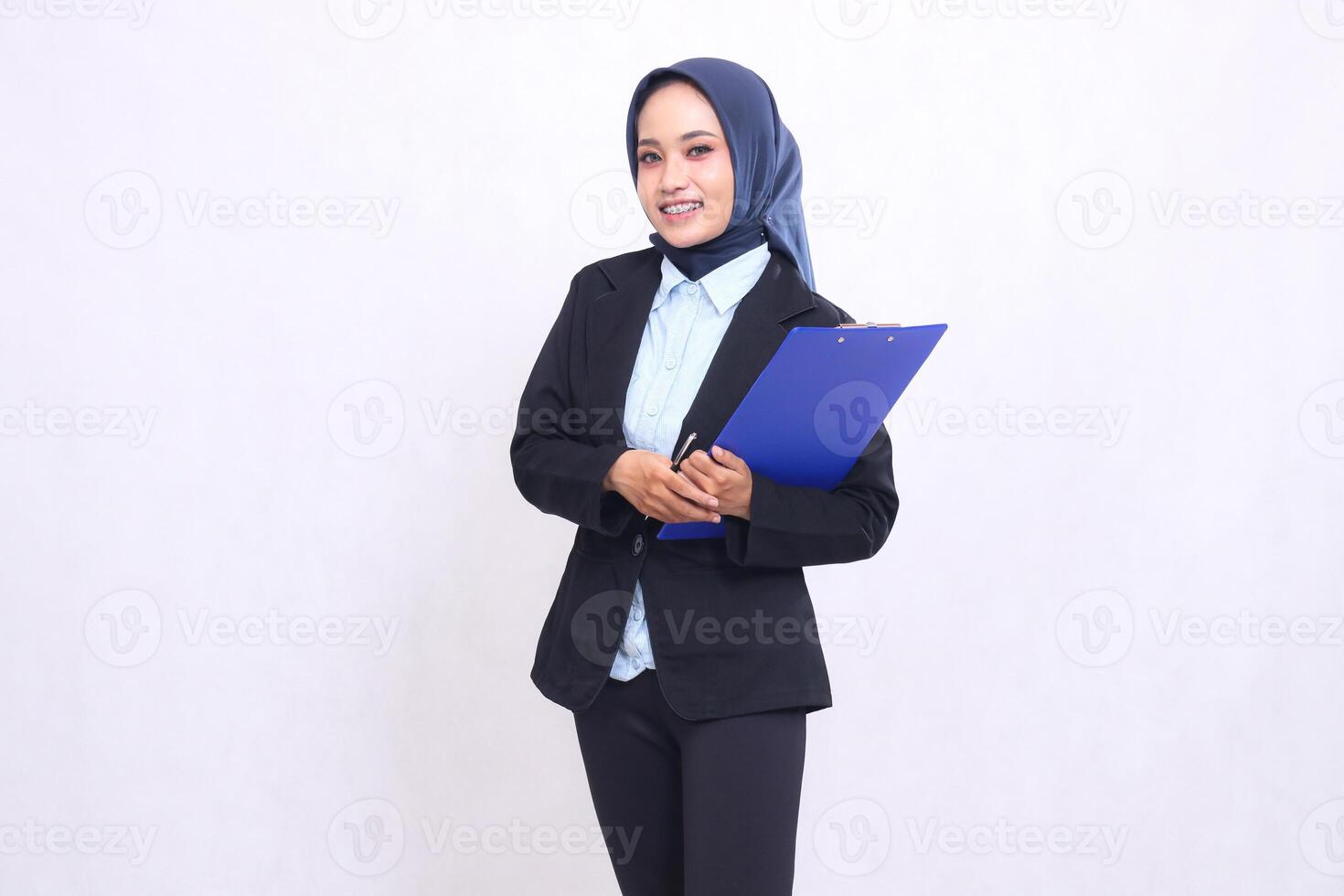 Mature Asian office woman wearing a hijab stands smiling cheerfully at the camera holding a pen and hugging a clipboard. Beautiful Muslim women wearing blue shirts for business, finance and corporate photo