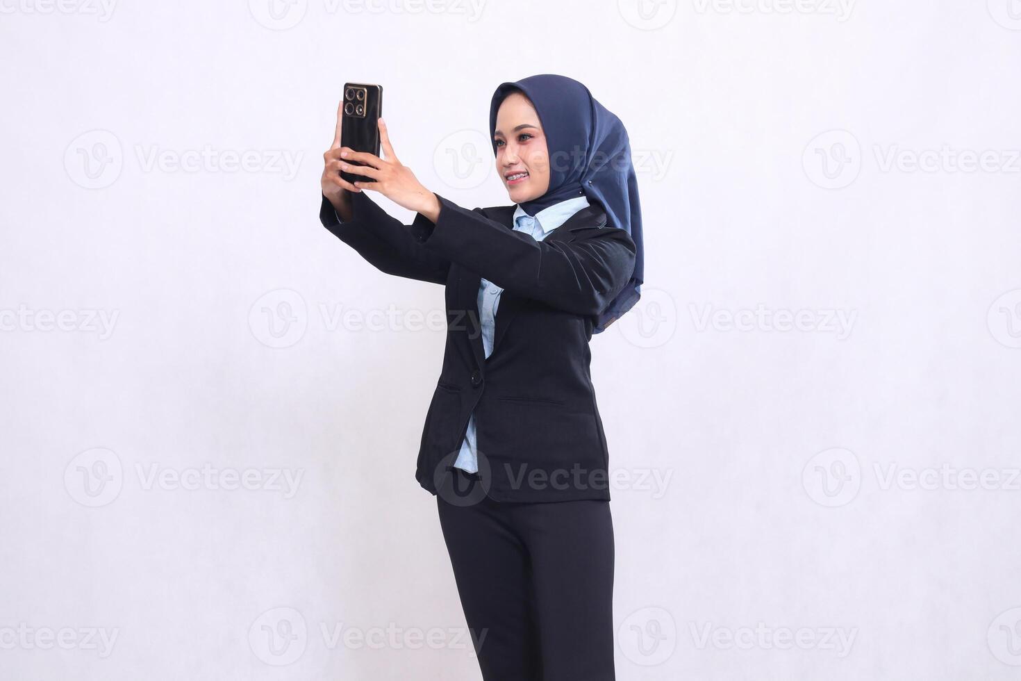 Young Asian office woman wearing a hijab stands cheerfully looking and holding her cell phone gadget up while taking a selfie. Beautiful Muslim woman in blue shirt used for finance and technology photo