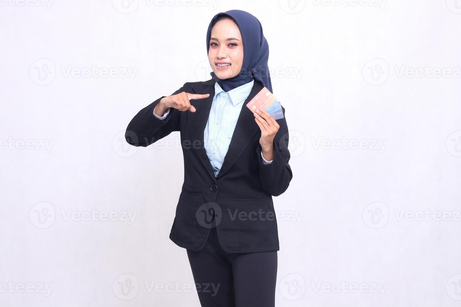 Asian mature office girl in hijab stands cheerfully pointing and carrying e-credit debit card above for easy digital transactions. adult Muslim blue shirt for finances, transactions and loans photo