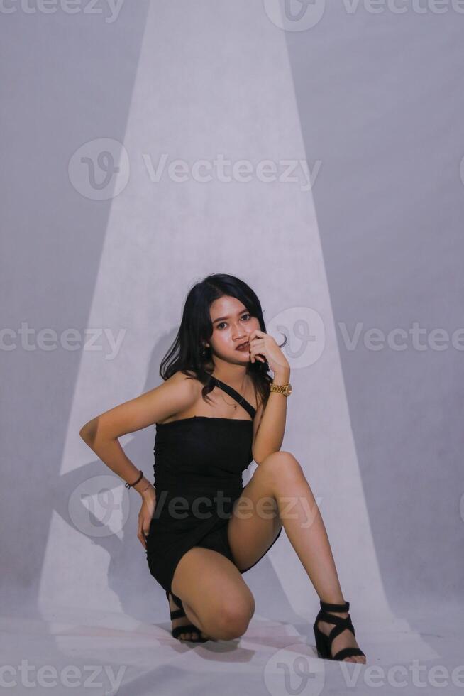 full body model Sexy elegant young Asian woman squatting with hands holding chin and buttocks wearing black mini dress with white background with spotlight photo