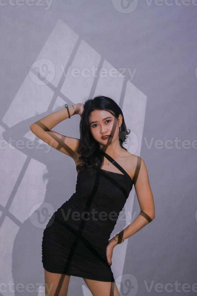 Sexy young Asian female model with hands holding her thighs and upper hair wearing a black mini dress on a white background with a window motif spotlight photo