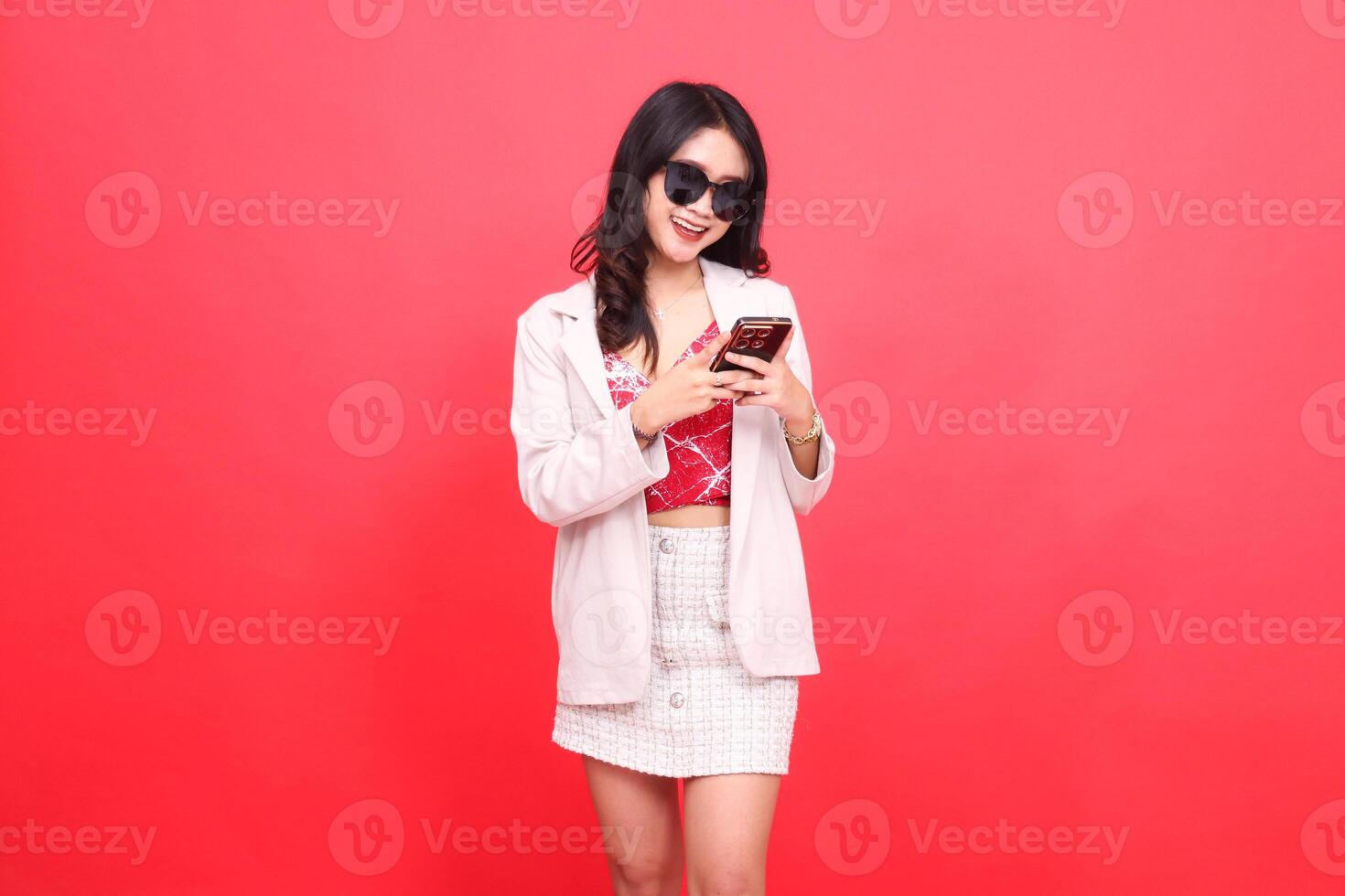 A beautiful Asian Indonesian woman wearing glasses, cheerfully slanted to the left, operates a cellphone cheerfully wearing a cream-colored blazer photo