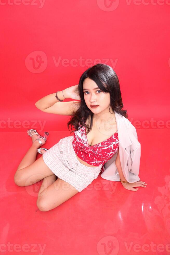 Sexy Asian Indonesian woman sitting crookedly at a high angle wearing a red patterned tank top and cream colored formal blazer, hands holding her hair photo