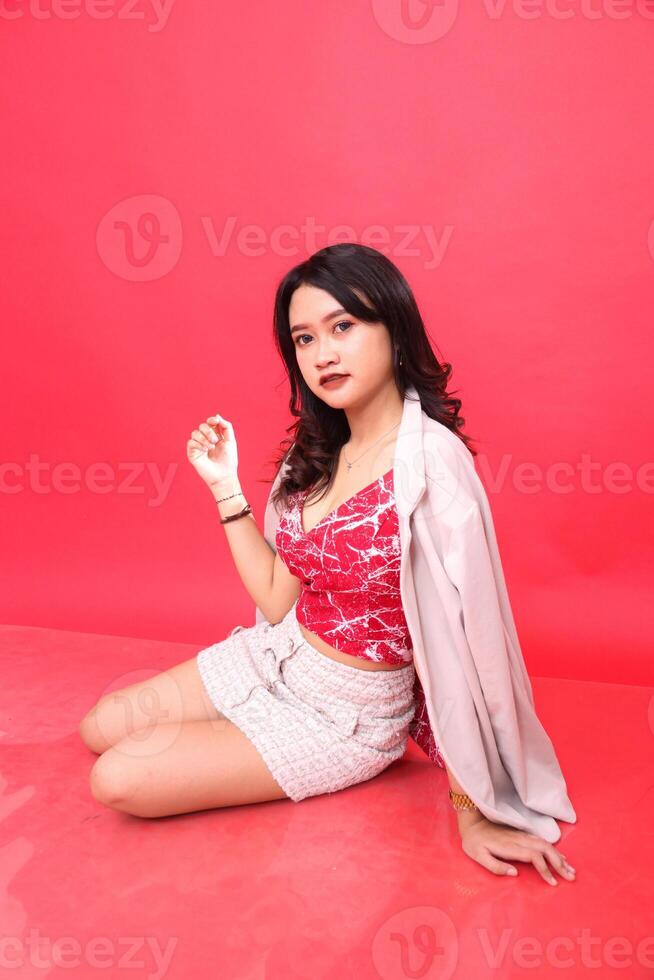 Sexy Asian Indonesian woman sitting crookedly upright wearing a red patterned tank top and cream colored formal blazer, hands posing as a model photo