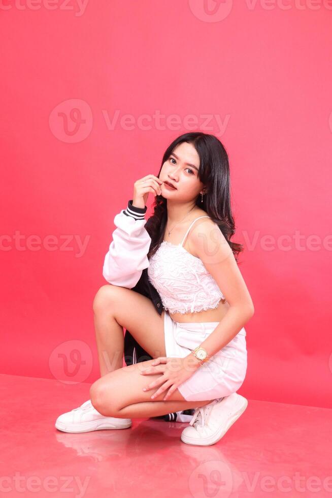 Beautiful young Asian woman wearing elegant sunglasses, hands holding her chin and thighs sitting on the floor wearing a black and white jacket photo