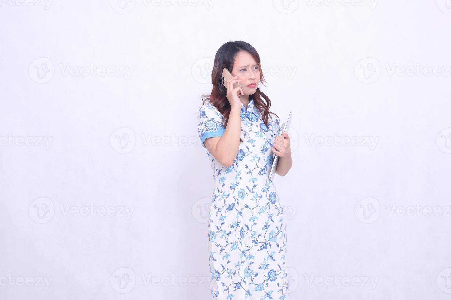beautiful Indonesian Chinese woman looking obliquely at top left wearing modern Chinese blue dress holding tablet while making phone call isolated on white background photo