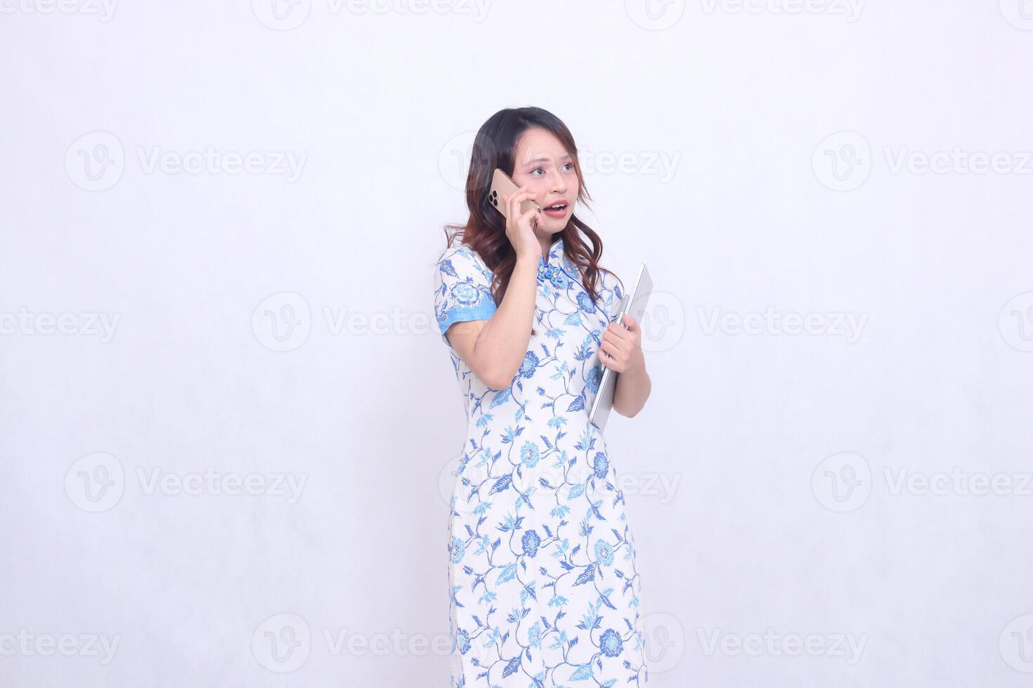 Beautiful Chinese Indonesian woman shocked looking at top left side wearing modern Chinese blue dress holding tablet while making phone call isolated on white background photo