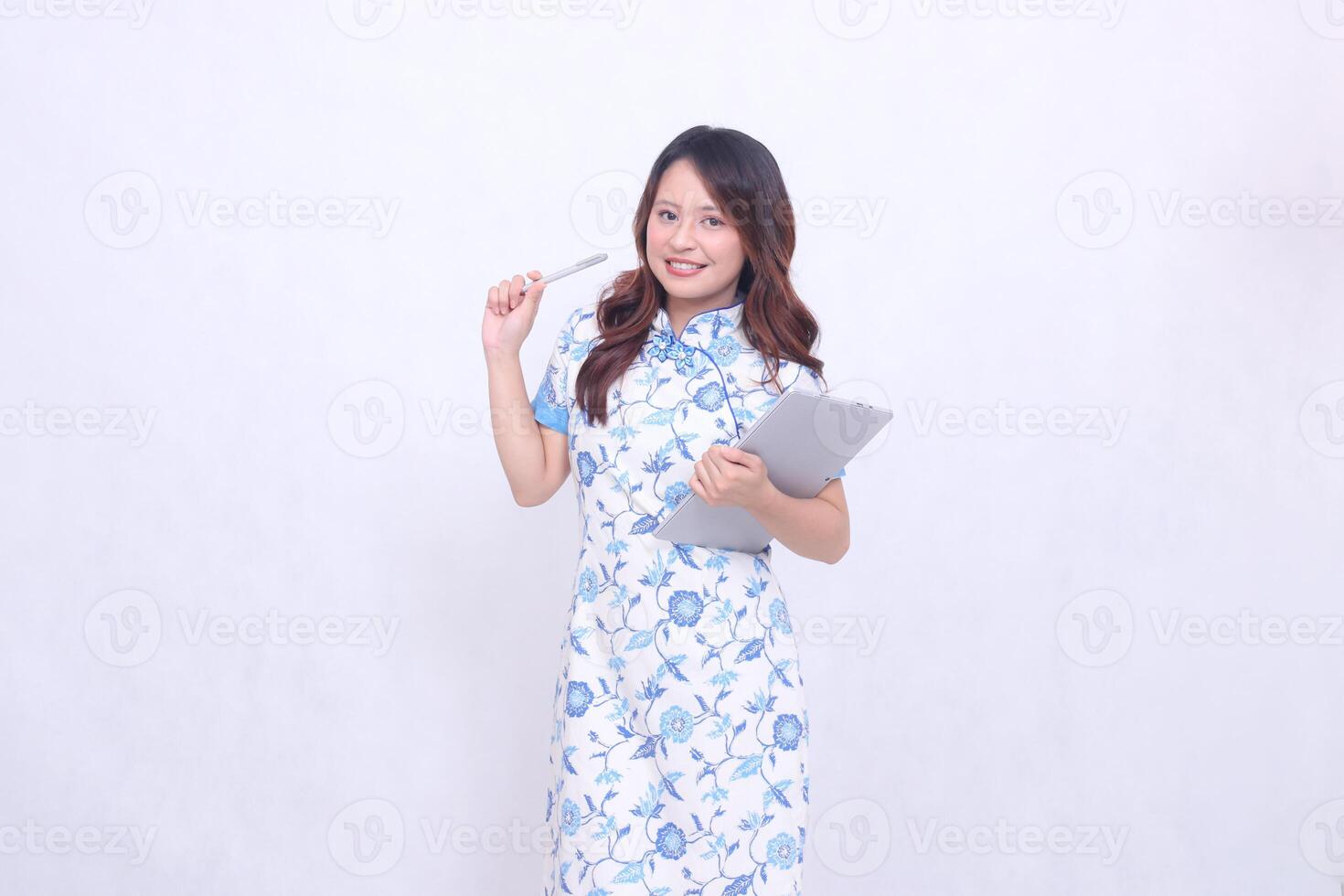 Beautiful cheerful Indonesian Chinese woman looking at camera wearing modern Chinese blue dress hugging tablet and pointing with pen isolated white background photo