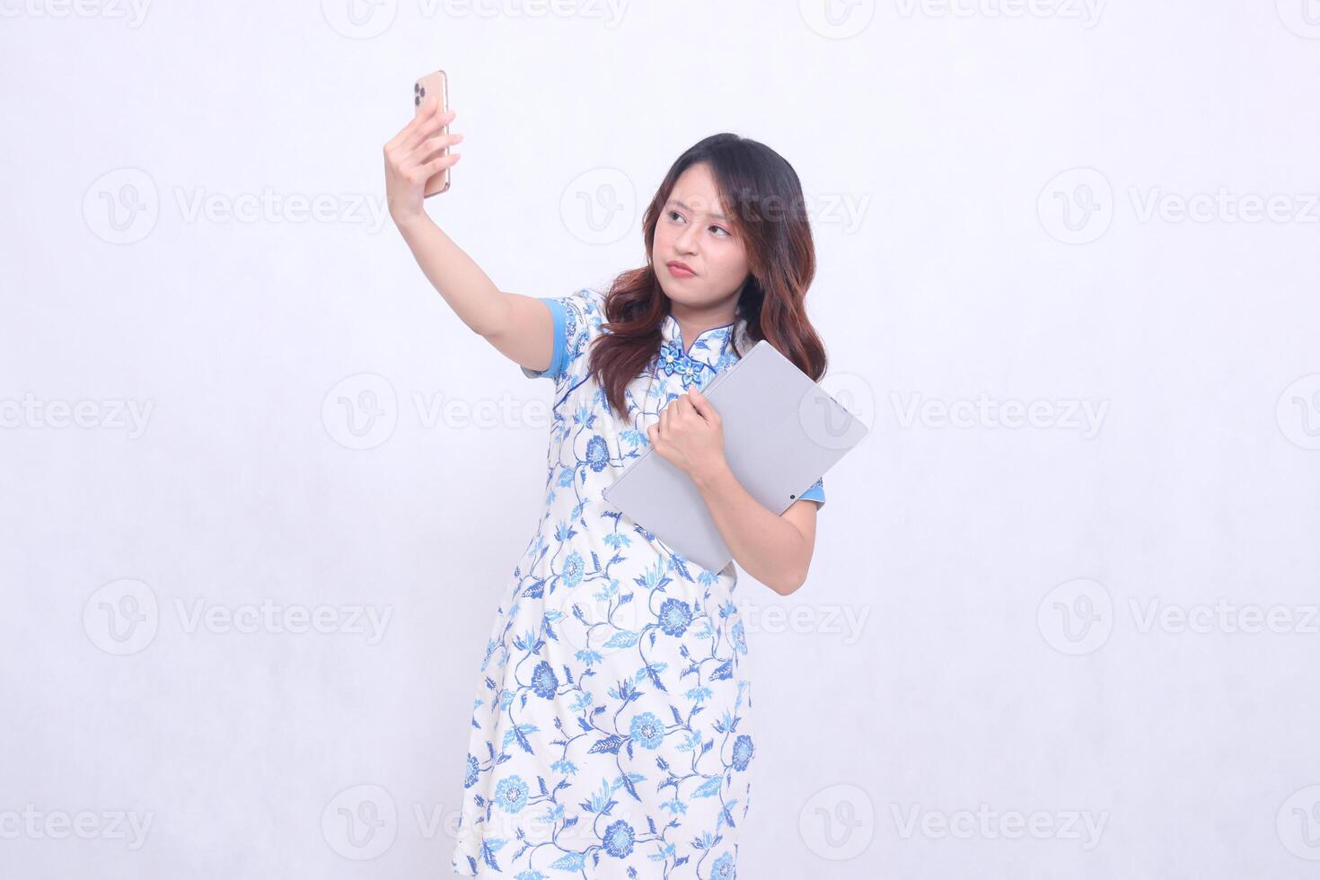 beautiful asian woman wearing modern chinese blue dress tilted to the right frowning holding tablet and looking at cell phone up isolated white background photo