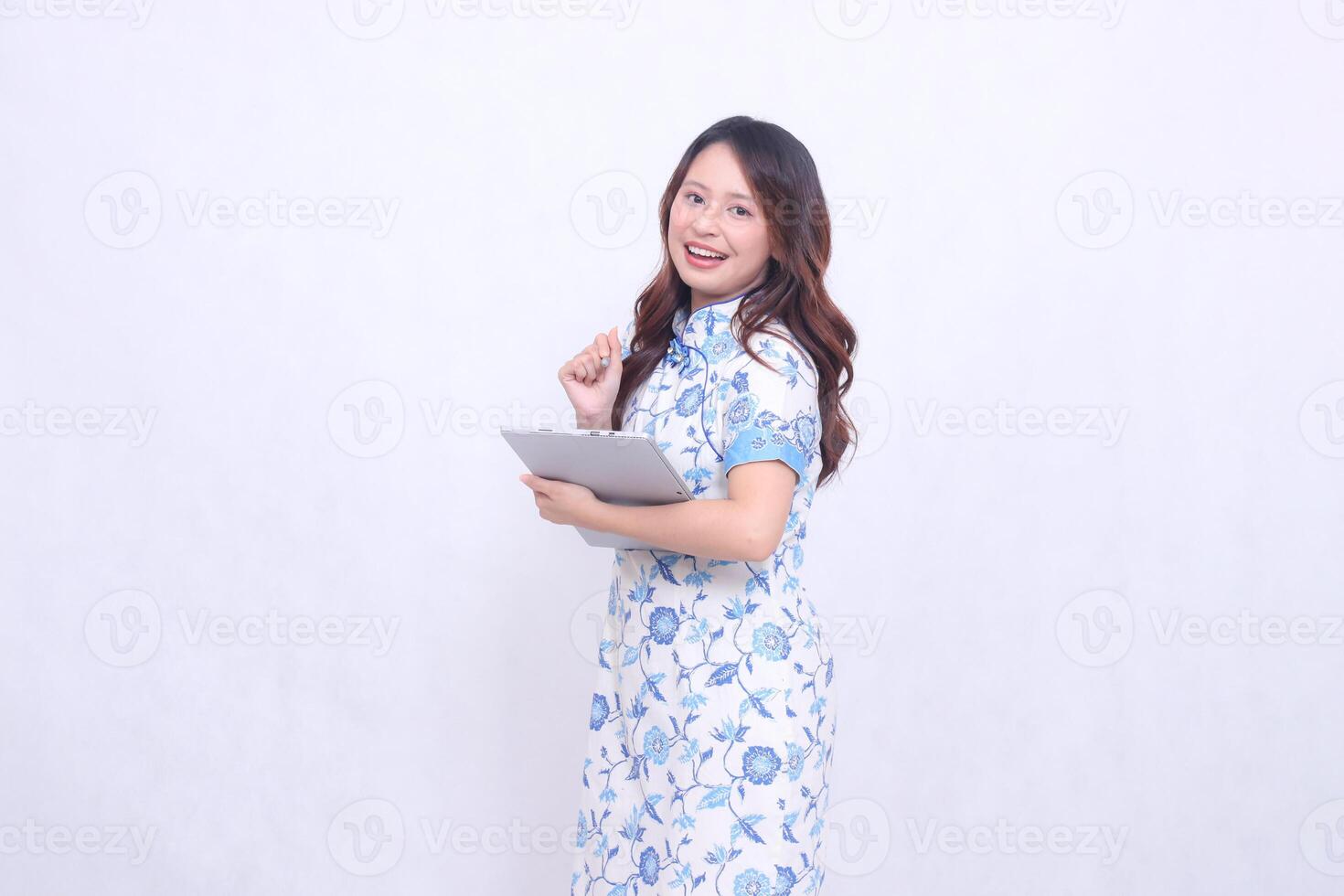 Beautiful cheerful Indonesian Chinese woman leaning to the right wearing a modern Chinese blue dress carrying a tablet, front view isolated on white background photo