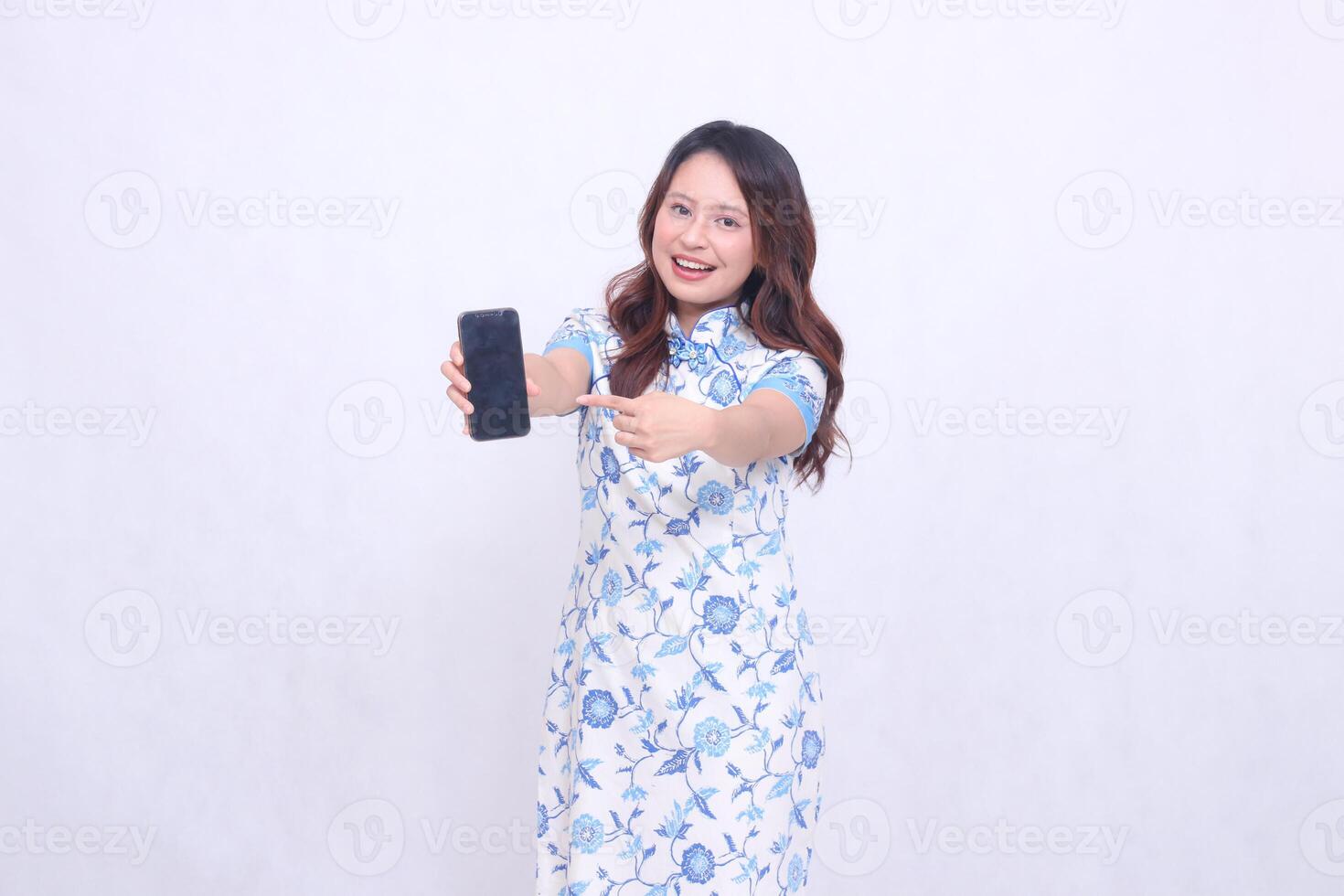 young Indonesian Javanese woman wearing blue traditional Chinese dress cheerful with arms wide holding gadget showing cellphone screen forward pointing to the side isolated white background photo