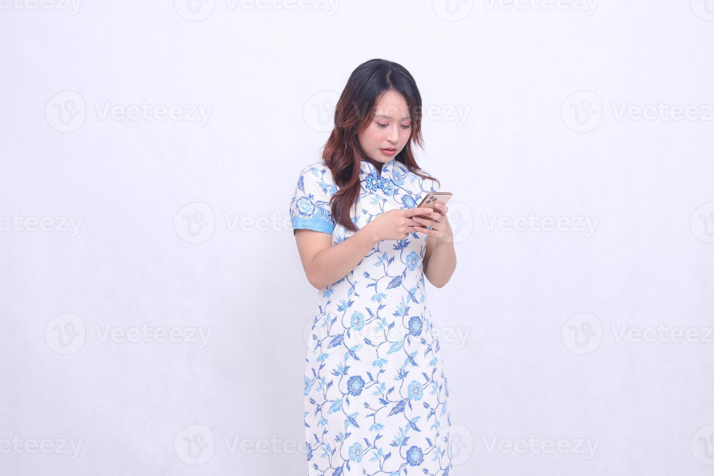 Young Asian Chinese Javanese woman wearing a blue Chinese dress is disappointed with her left side while focusing on typing on a gadget isolated on a white background photo
