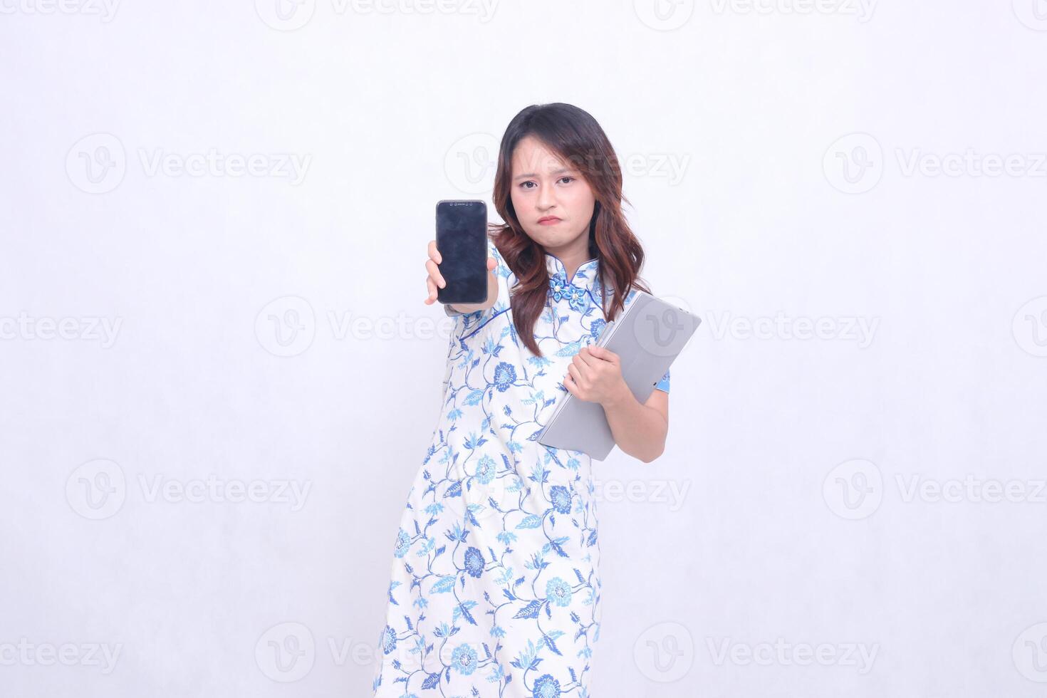 beautiful asian woman wearing blue chinese dress looking at camera frowning holding tablet and looking at cellphone screen straight isolated white background photo