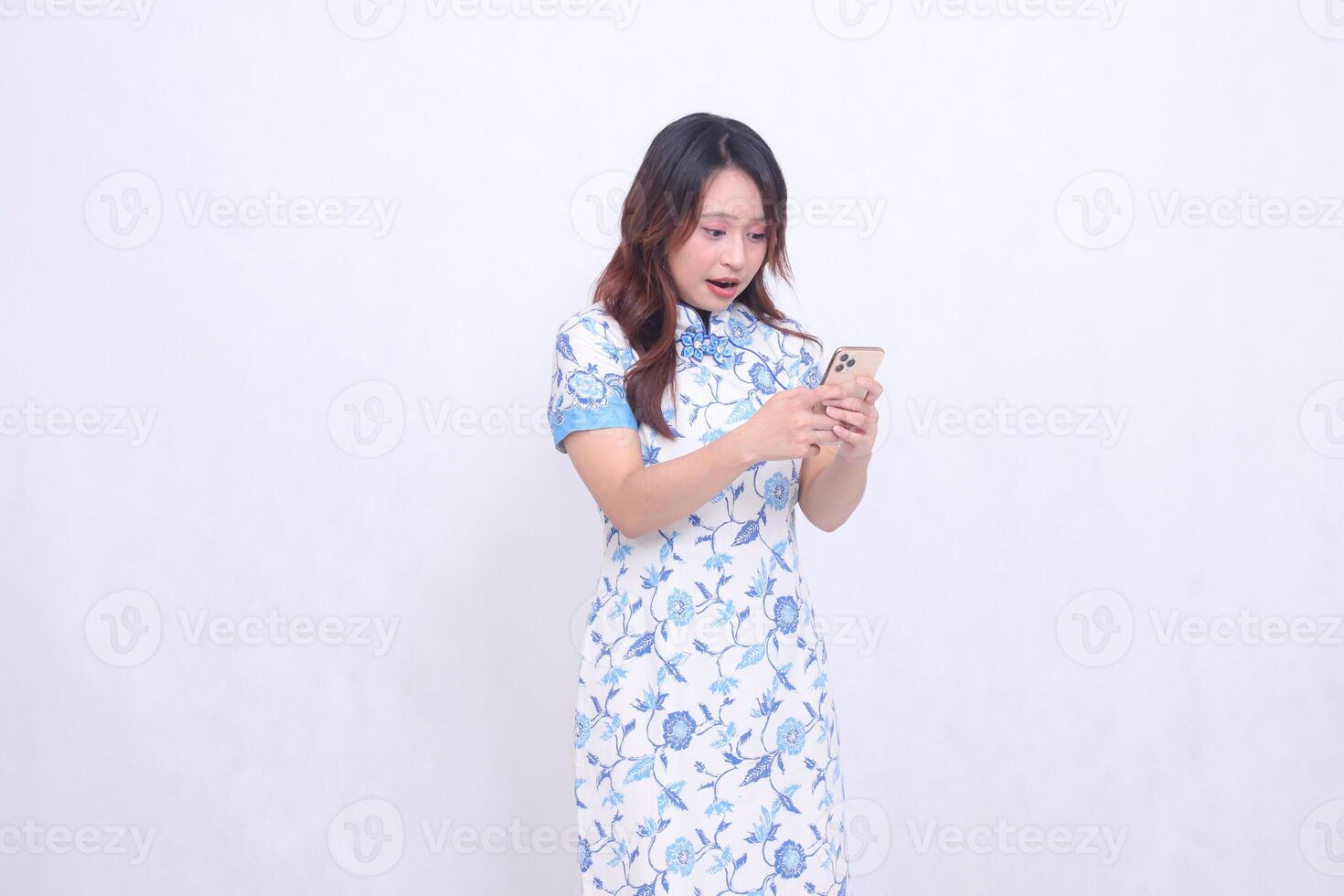 young Asian Javanese Chinese woman wearing a blue Chinese dress in shock, tilting to the left while both hands focus on typing on a cellphone isolated on a white background photo