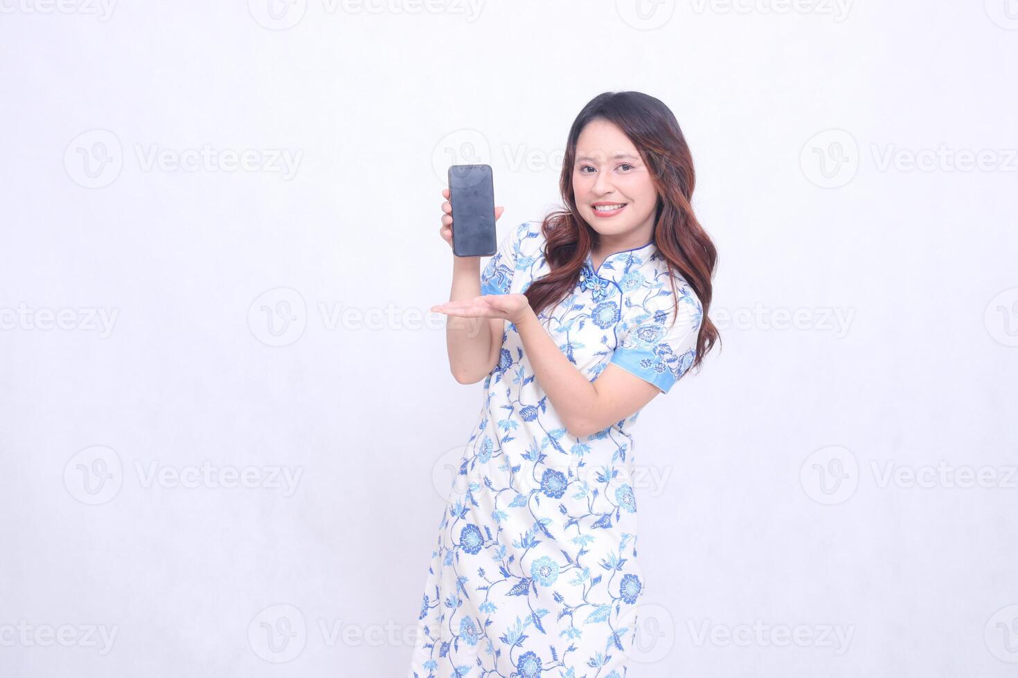 Young Asian Javanese Chinese girl wearing traditional Chinese blue dress cheerfully promoting gadgets on cell phone screen isolated white background photo