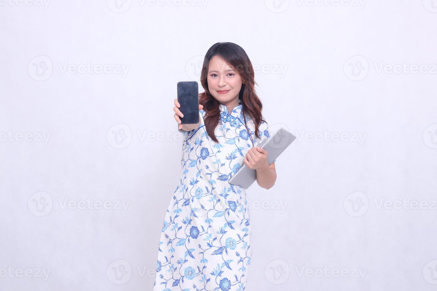 Beautiful Asian woman wearing a blue Chinese dress looks at the camera, smiles faintly, holds a tablet and looks at the cellphone screen, standing tall, isolated on a white background photo