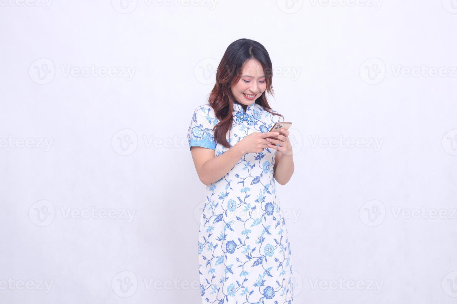young Asian Chinese Javanese woman wearing a blue Chinese dress is happy leaning to the left while focusing on typing on a gadget isolated on a white background photo