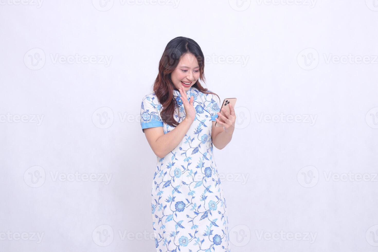 Young Asian Javanese Chinese woman smiles wearing a left-slanted Chinese blue dress while operating a gadget saying hello to communicating isolated on a white background photo
