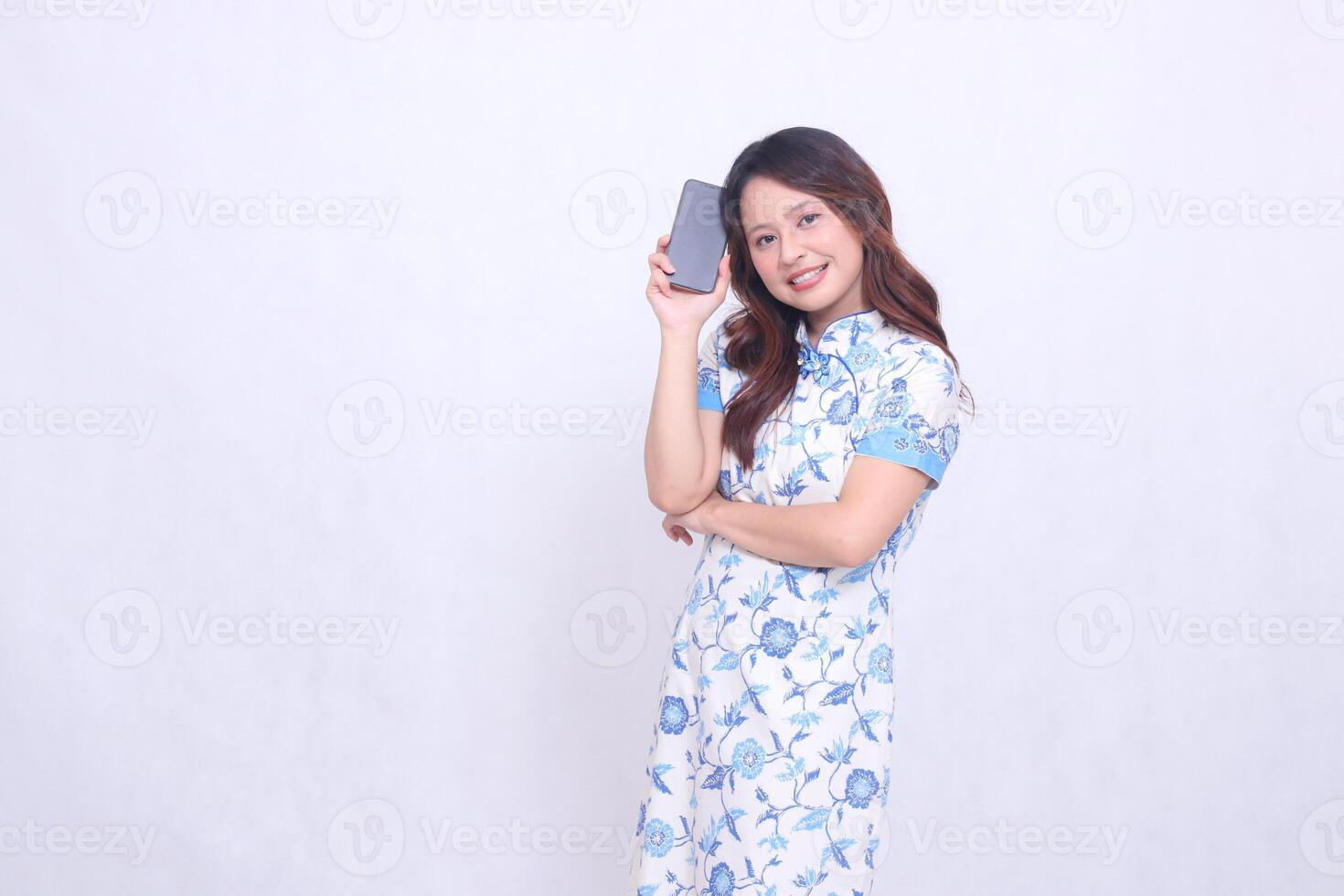 young Indonesian Javanese woman wearing a blue traditional Chinese dress cheerful with her arms holding a gadget showing an upright cellphone screen isolated on a white background photo