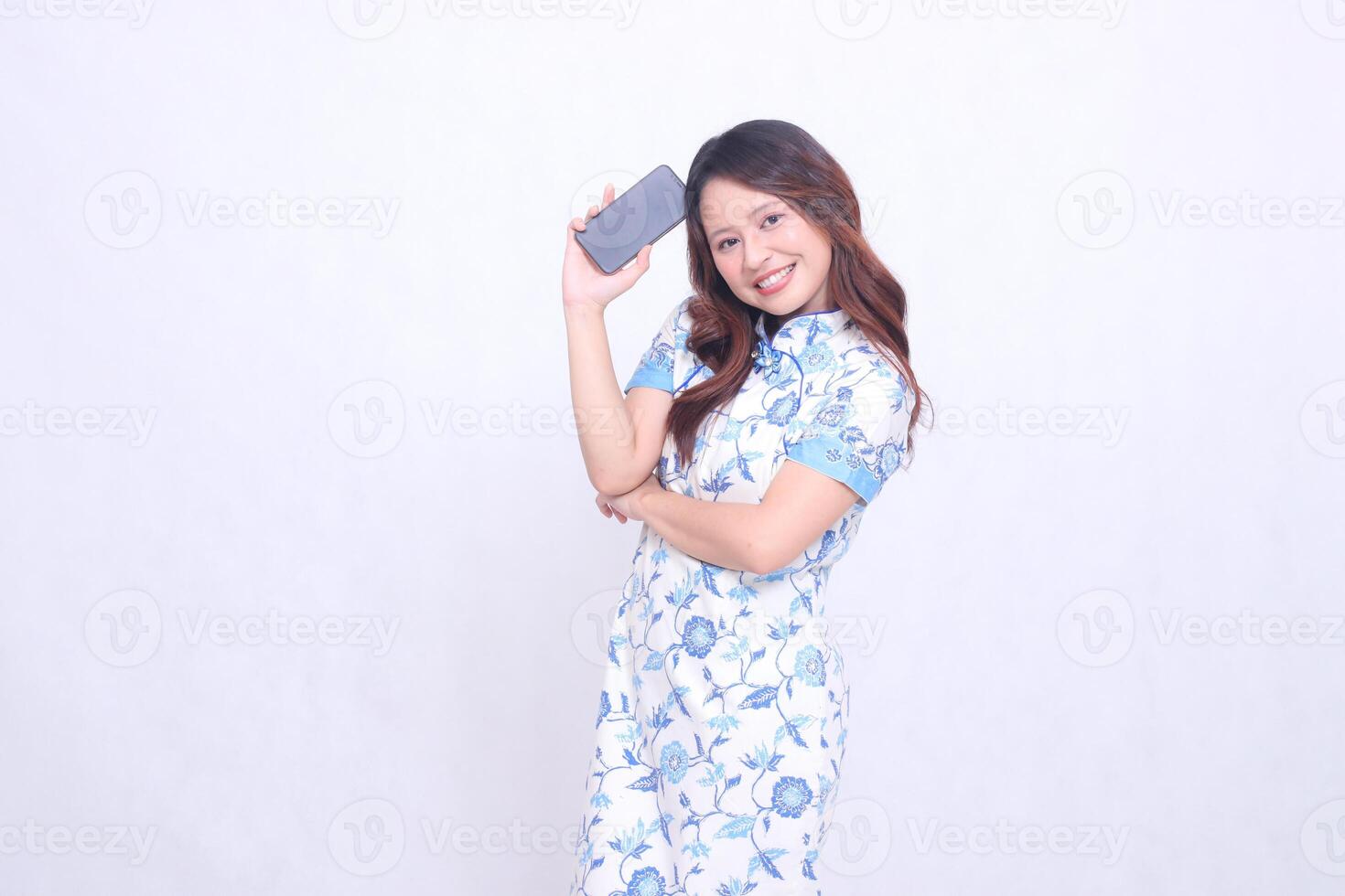 young Indonesian Javanese woman wearing blue traditional Chinese dress happy holding gadget showing tilted cell phone screen isolated white background photo