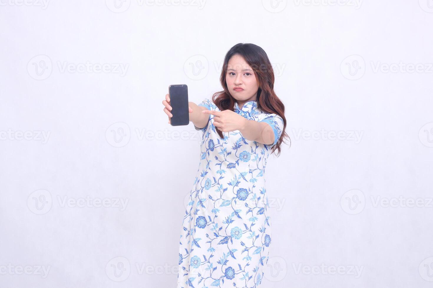 young Indonesian Javanese woman wearing blue traditional Chinese dress frowning holding gadget wide showing cell phone screen forward pointing to the side isolated white background photo