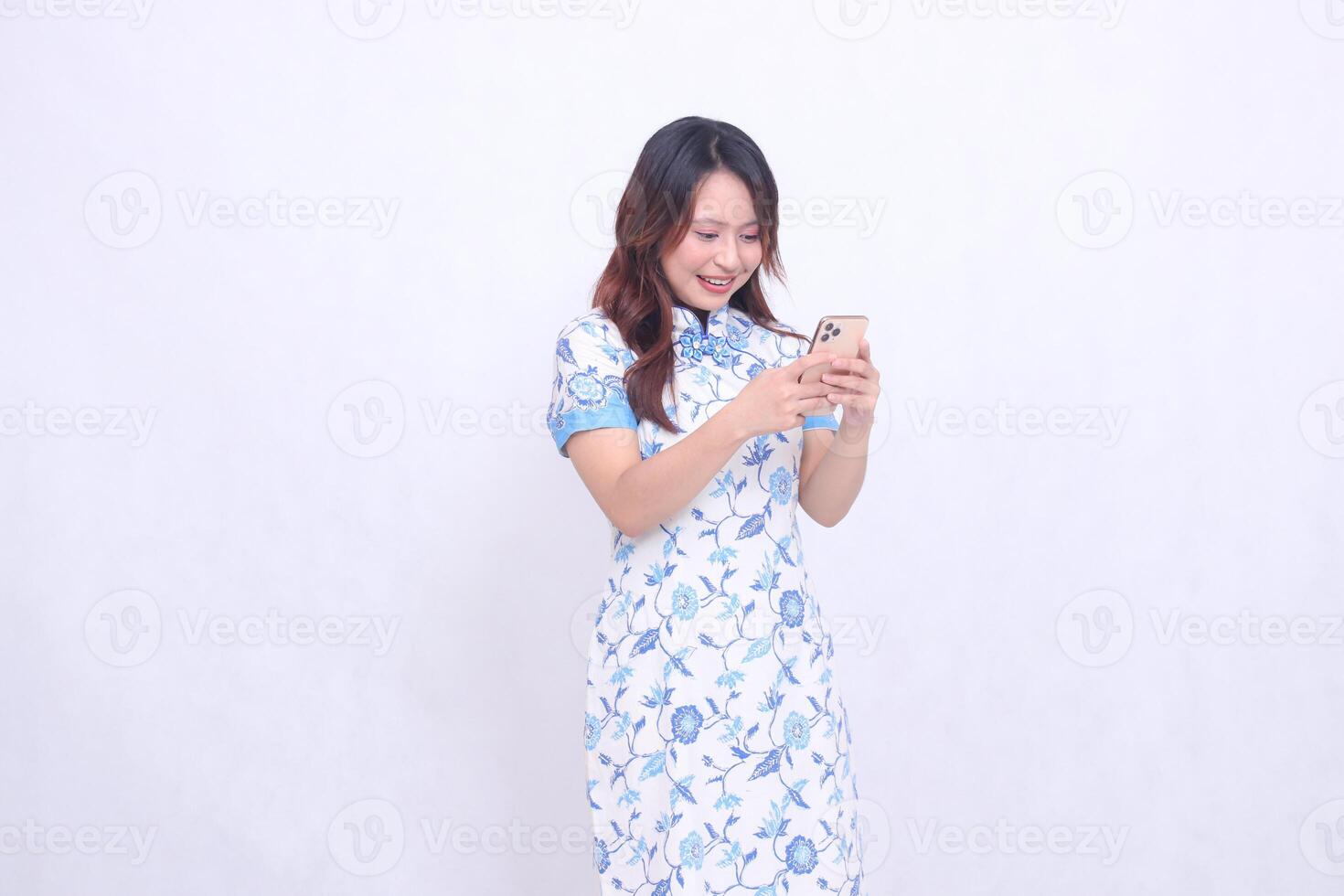 Young Asian Chinese Javanese woman wearing a blue Chinese dress is happy on her left side while both hands are focused on typing on her cellphone isolated on a white background photo